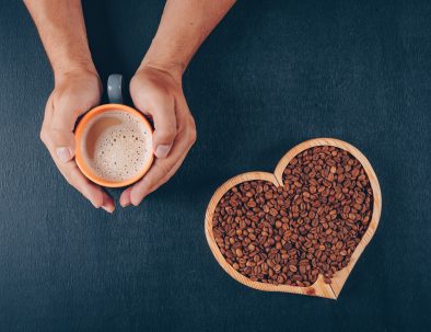 Man holding coffee with coffee beans in a heart shaped bowl on black background, top view.